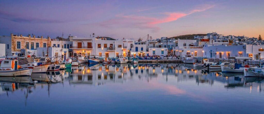 Evening view of Parikia harbor with boats and lit buildings reflecting on the water.