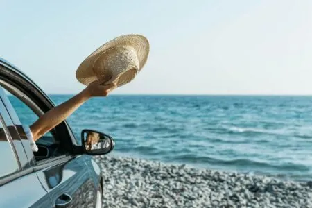 Woman holding a straw hat out of a car window near the sea in Paros, Greece.