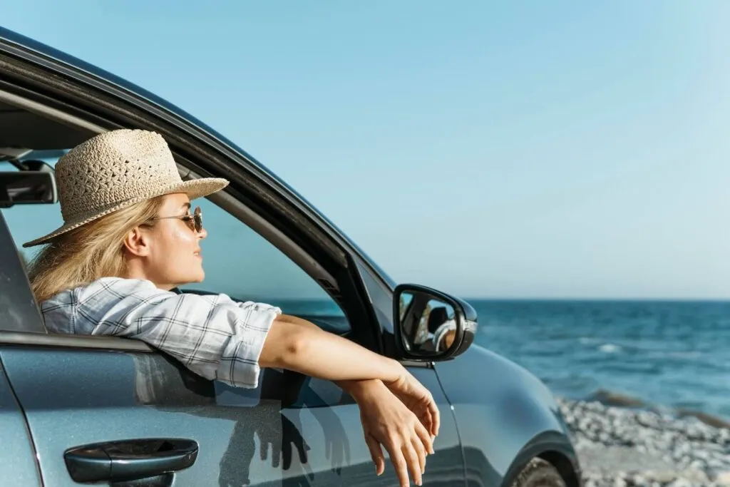 A woman wearing a straw hat and sunglasses leans out of a car window, smiling and enjoying the view of the sea on Paros Island. The image highlights a peaceful moment during a "rent a car" journey along the coastline.